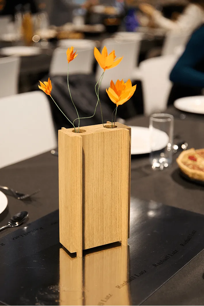 Une table moderne est dressée avec un vase en bois accueillant de délicates fleurs orange. En arrière-plan, des éléments de table flous créent une ambiance chaleureuse.