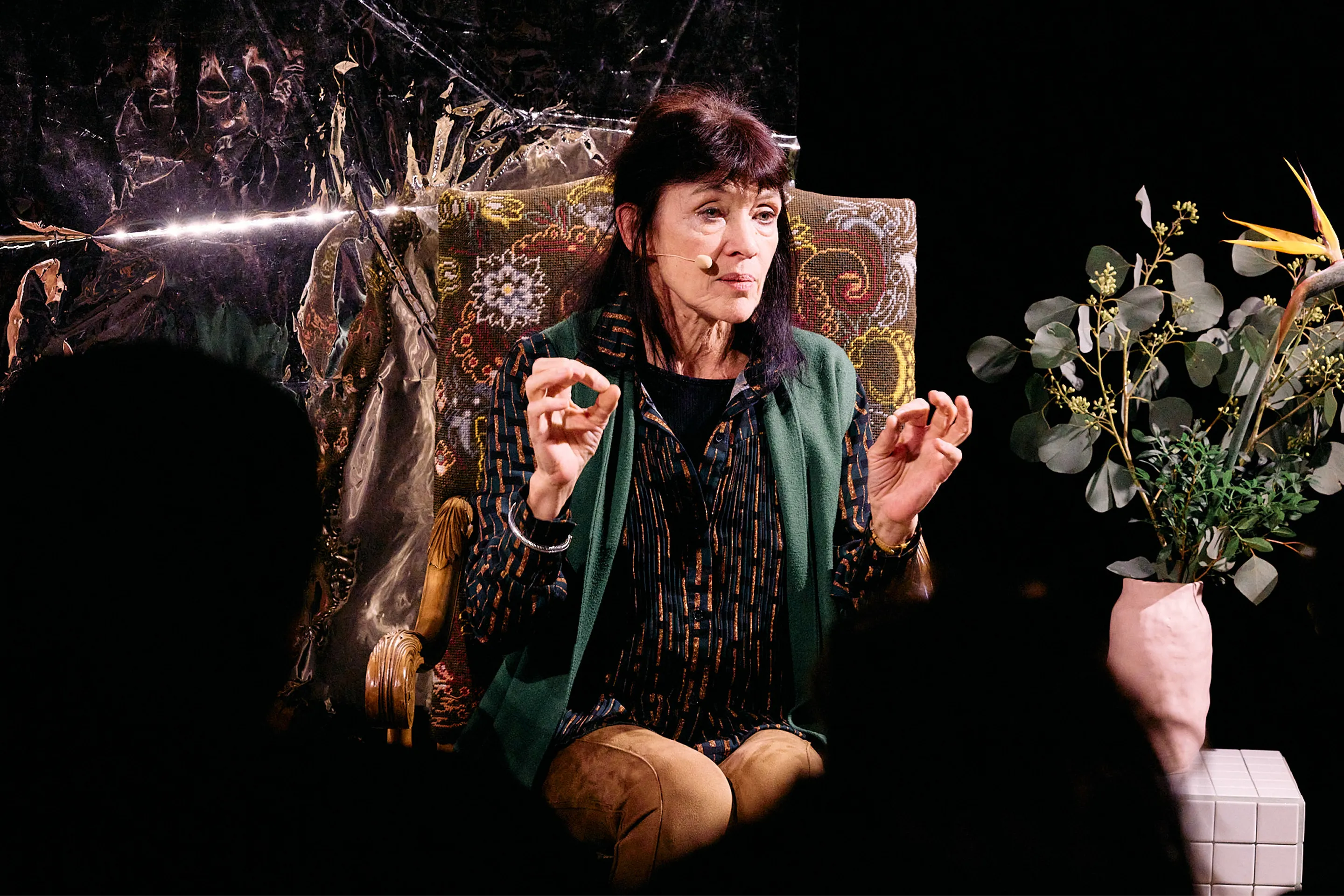 Femme assise sur un fauteuil orné, parlant avec un microphone, dans un cadre théâtral avec des éléments artistiques en arrière-plan et un vase de fleurs à côté.